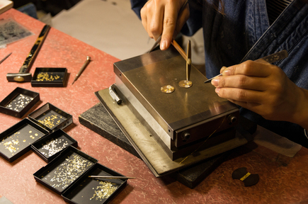 A Japanese artisan delicately inlays fine gold patterns onto a circular metal plate using traditional tools in the intricate process of Kyo-zogan (Kyoto metal inlay craftsmanship).